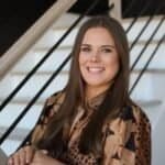 A woman with long brown hair, wearing a brown patterned blouse, smiles while standing in front of a staircase with black railings.
