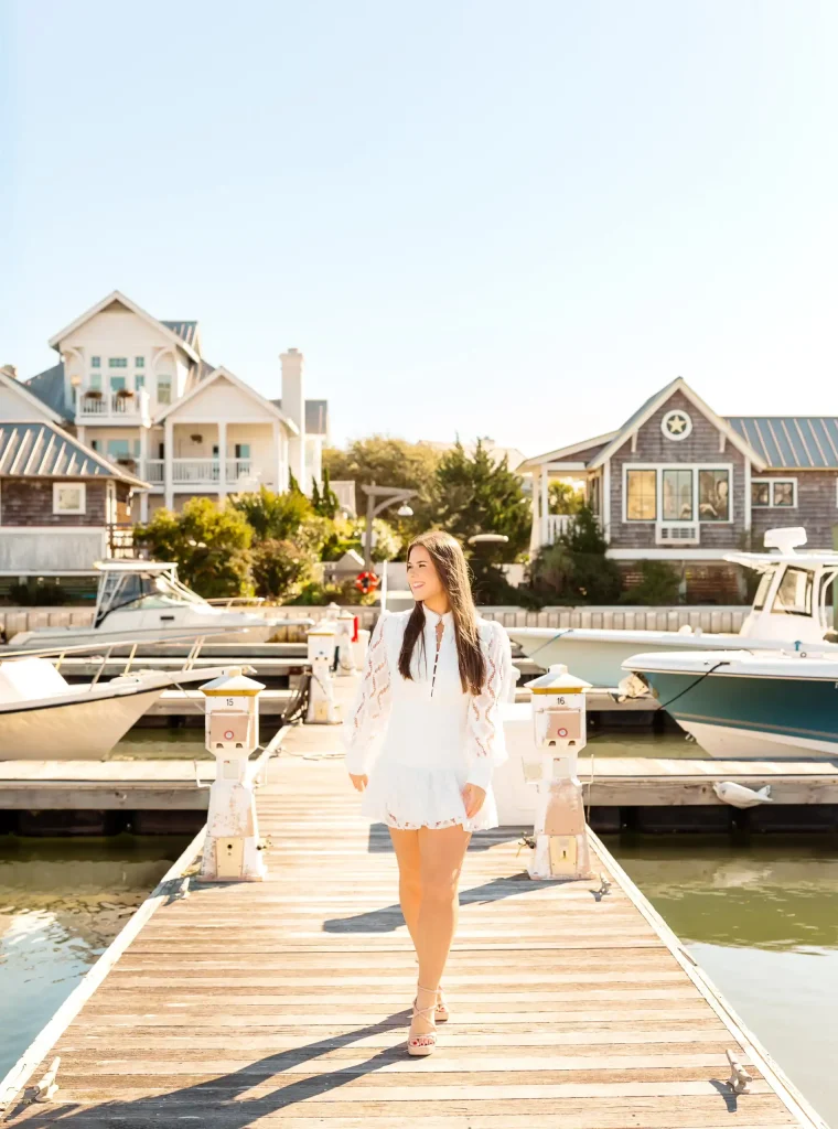 A woman in a white dress walks on a dock lined with boats, with charming coastal houses and bright sunlight in the background.