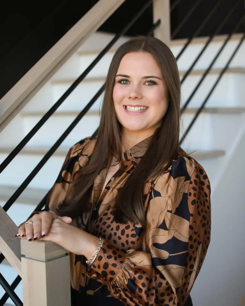 A woman with long brown hair smiles at the camera while standing by a staircase. She is wearing a brown and black patterned blouse and has her hands resting on a wooden railing.